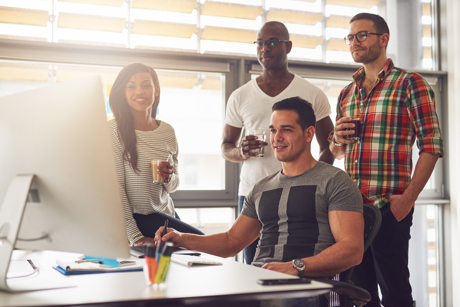 Four relaxed young diverse male and female small business workers with drinks in hand standing around computer in office